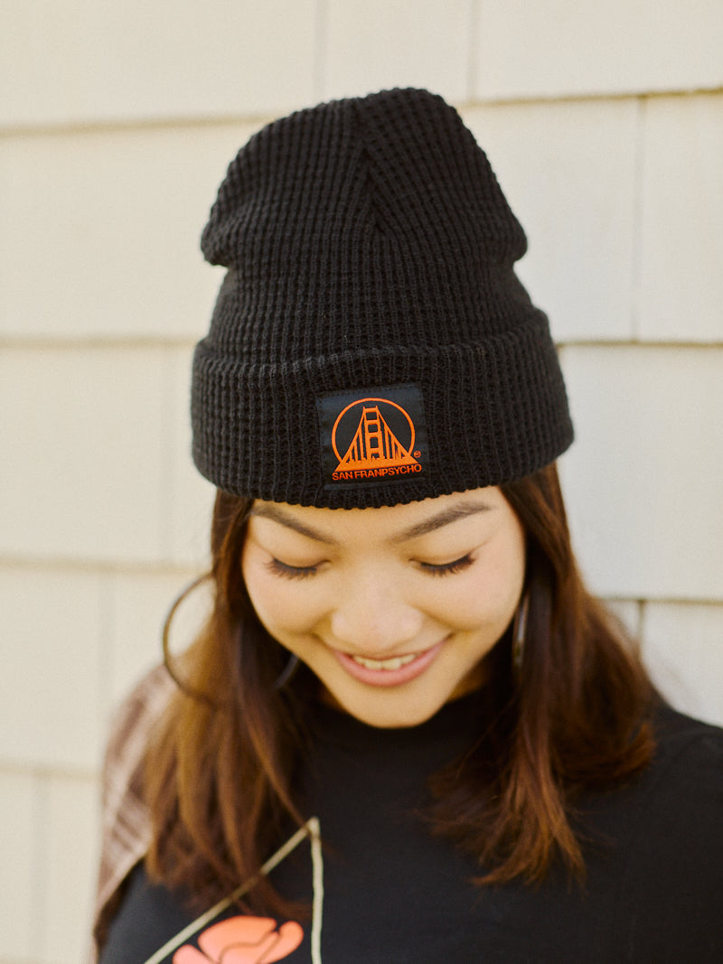 Woman wearing a black waffle beanie with a black/red San Franpsycho logo, looking down and smiling in front of a light wall.