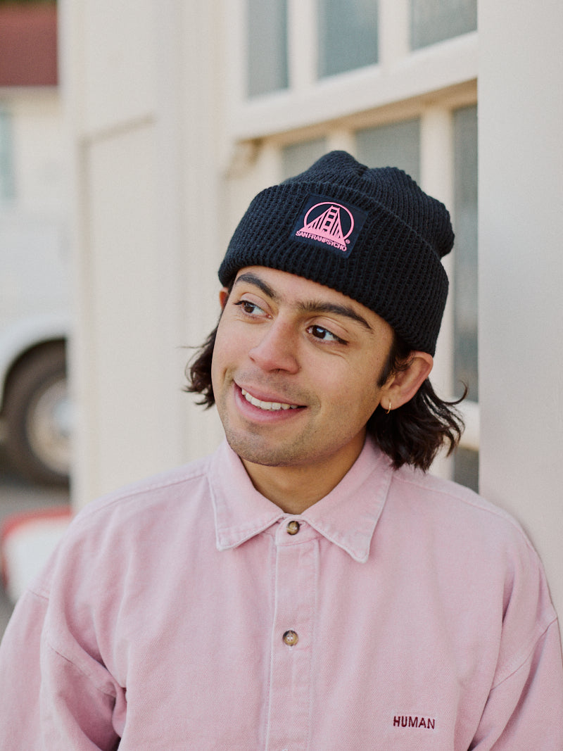 A person in a pink shirt and the Black Waffle Beanie with Black/Pink SFP Logo from San Franpsycho, smiling as they lean against a white wall outdoors.