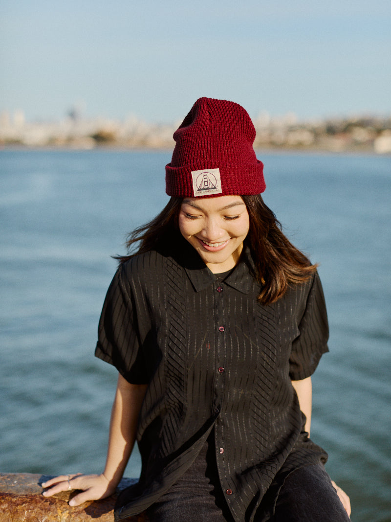 Woman in a Burgundy Waffle Beanie w/ Natural/Black SFP Logo and black shirt smiles by the water with a city skyline in the background.